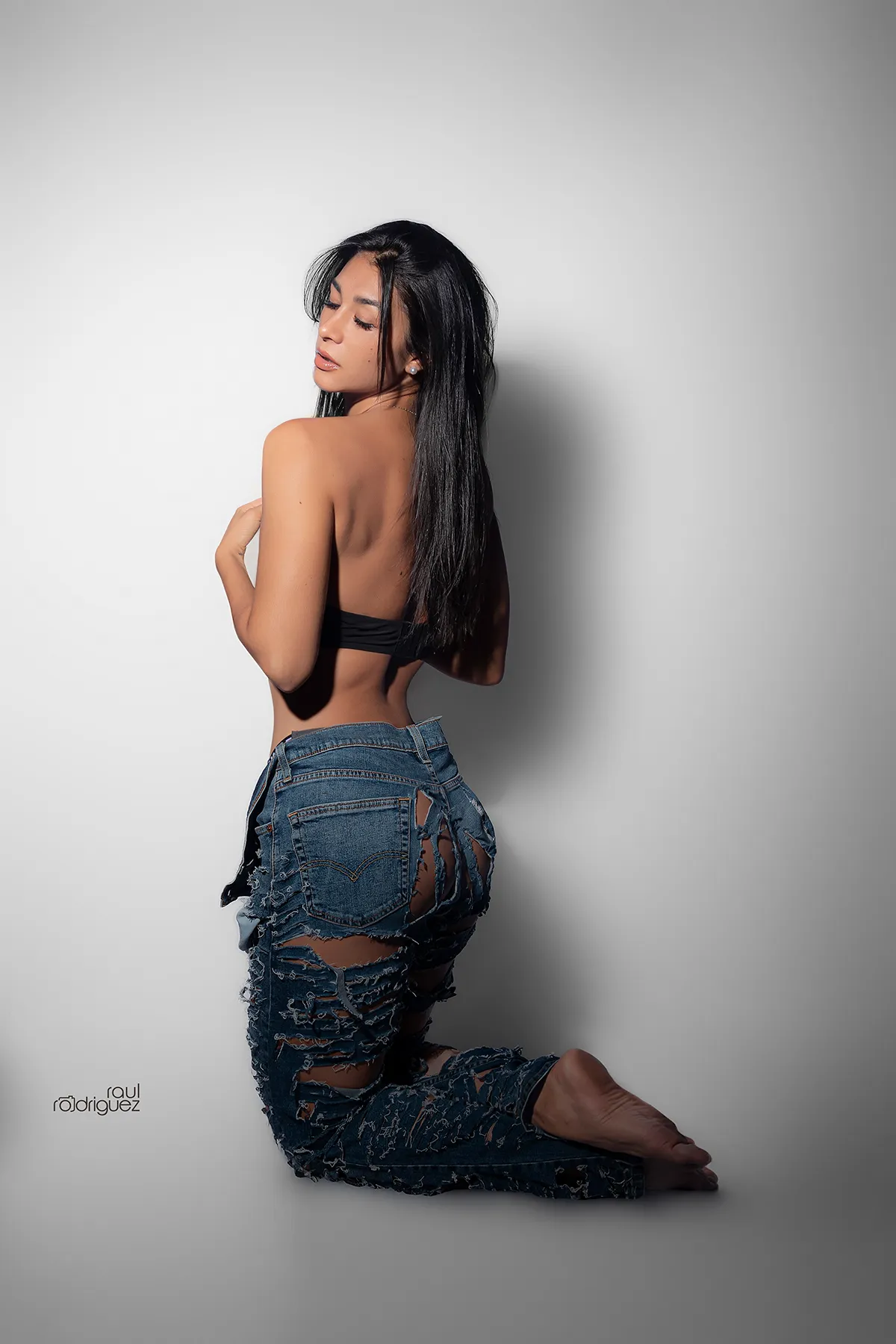 Mujer arrodillada de espaldas con top negro y jeans azules desgarrados, que gira el rostro hacia un lado sobre un fondo gris claro.