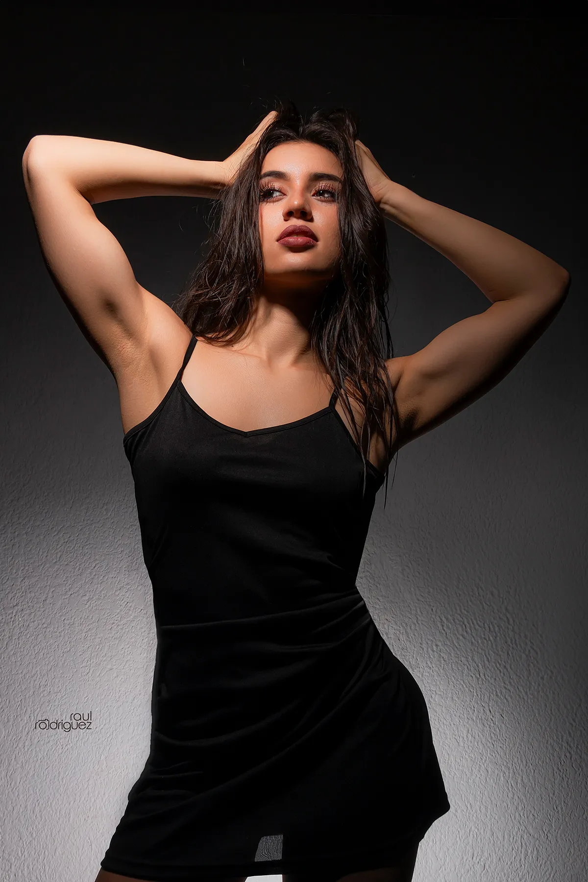 Retrato frontal de mujer con vestido negro y ambos brazos elevados, mirando hacia un lado bajo iluminación dura.