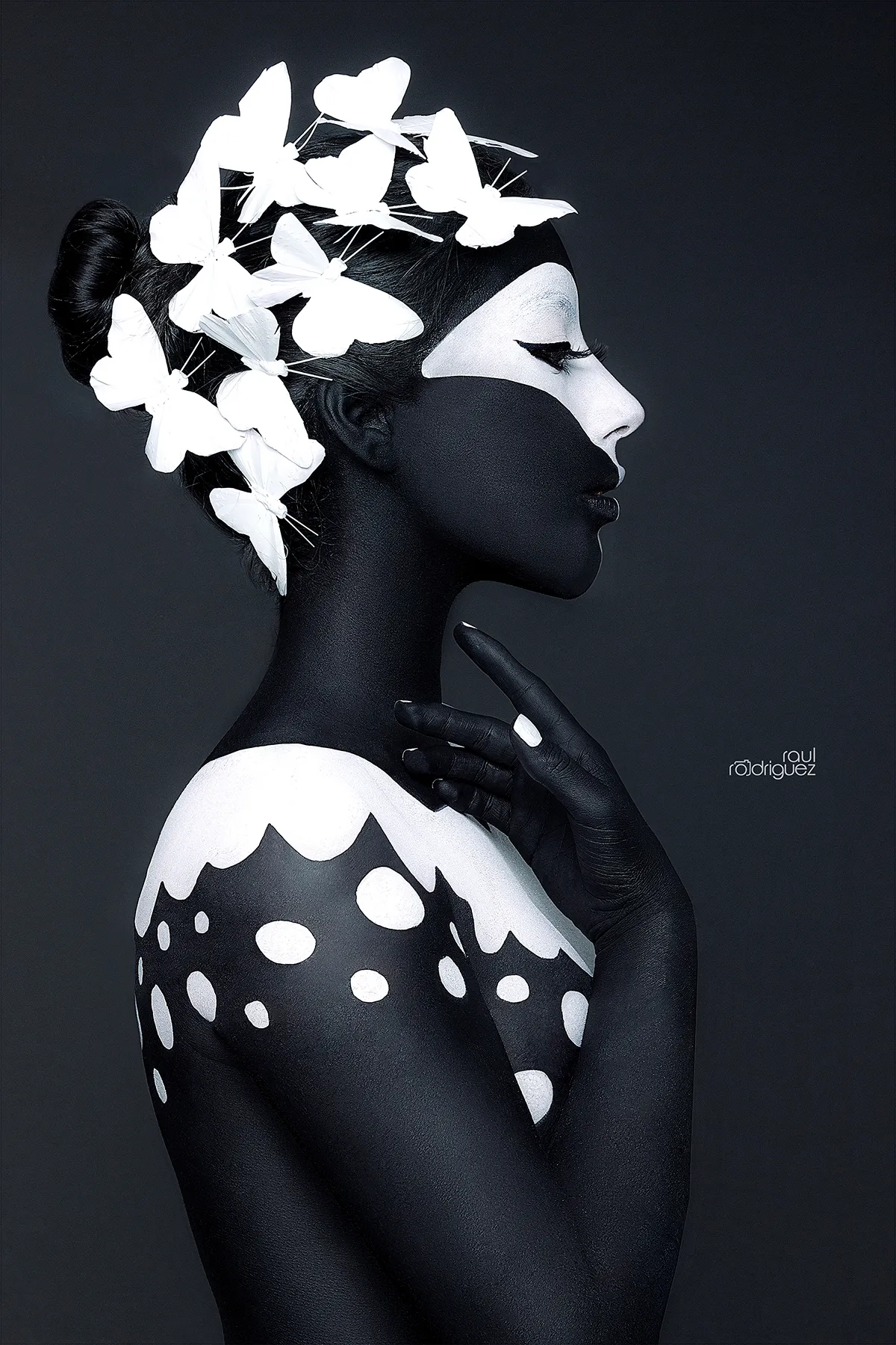 Retrato de perfil con maquillaje negro y blanco, mariposas blancas en el cabello y una mano elevada junto al cuello.