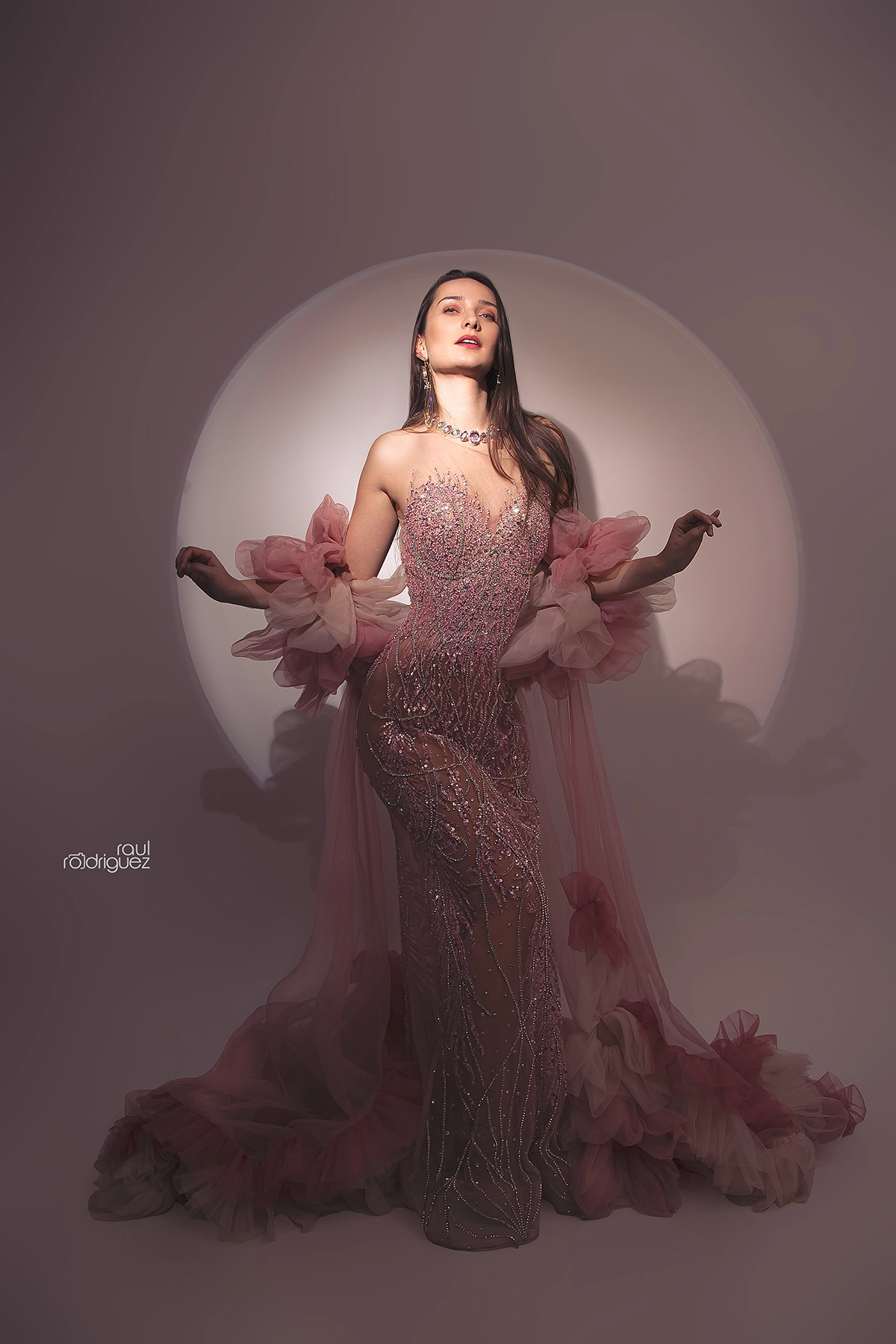 Figura de cuerpo completo con vestido rosa de pedrería y volúmenes de tul, frente a un círculo de luz.
