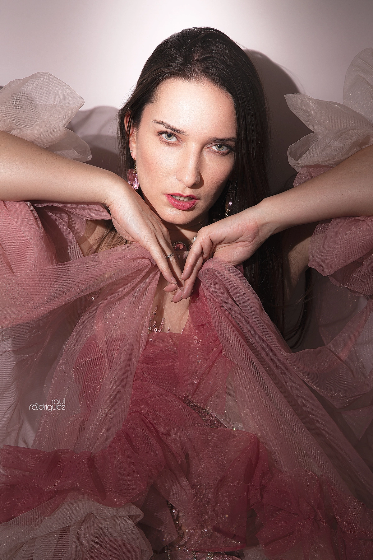 Retrato cerrado con mirada directa, manos bajo el rostro y tul rosa rodeando la figura.