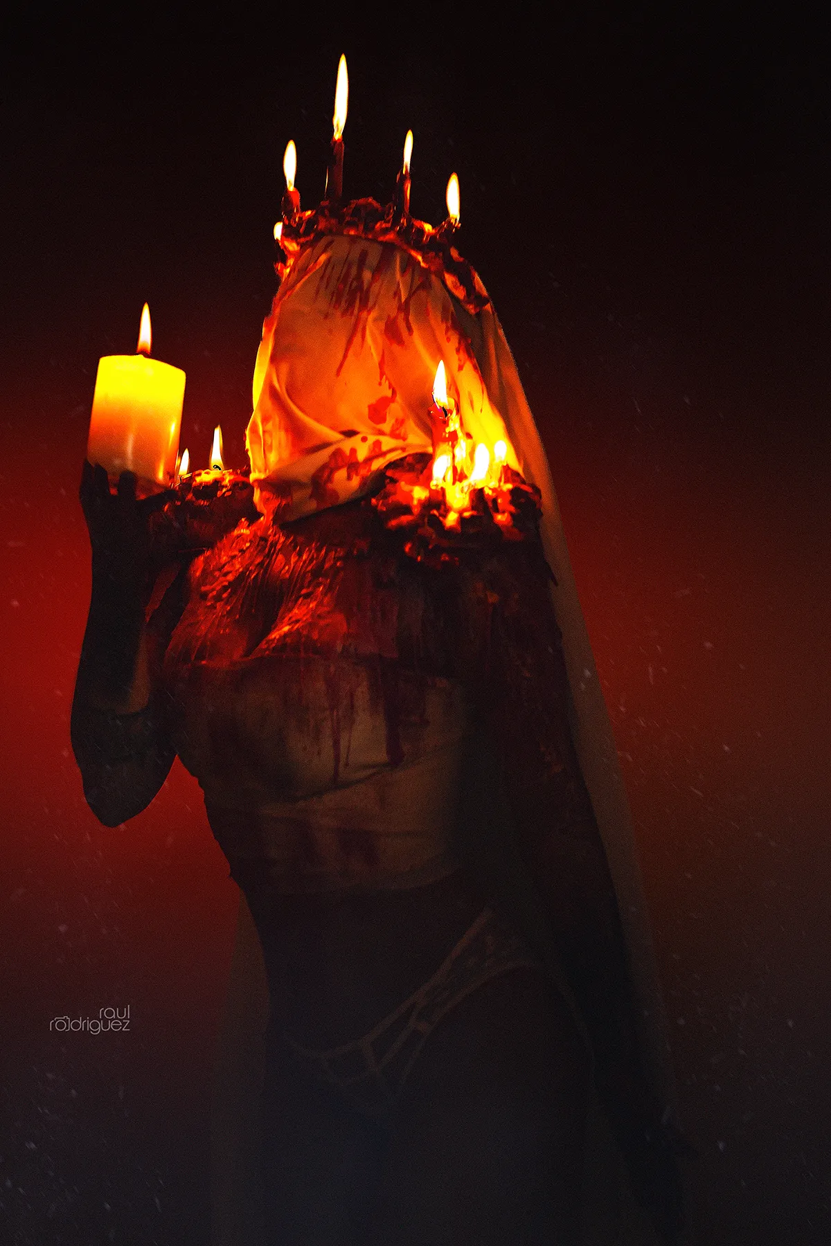 Figura de tres cuartos con el rostro cubierto por una tela clara con marcas rojizas, velas encendidas en la cabeza y los hombros y una vela gruesa elevada a un lado del rostro.