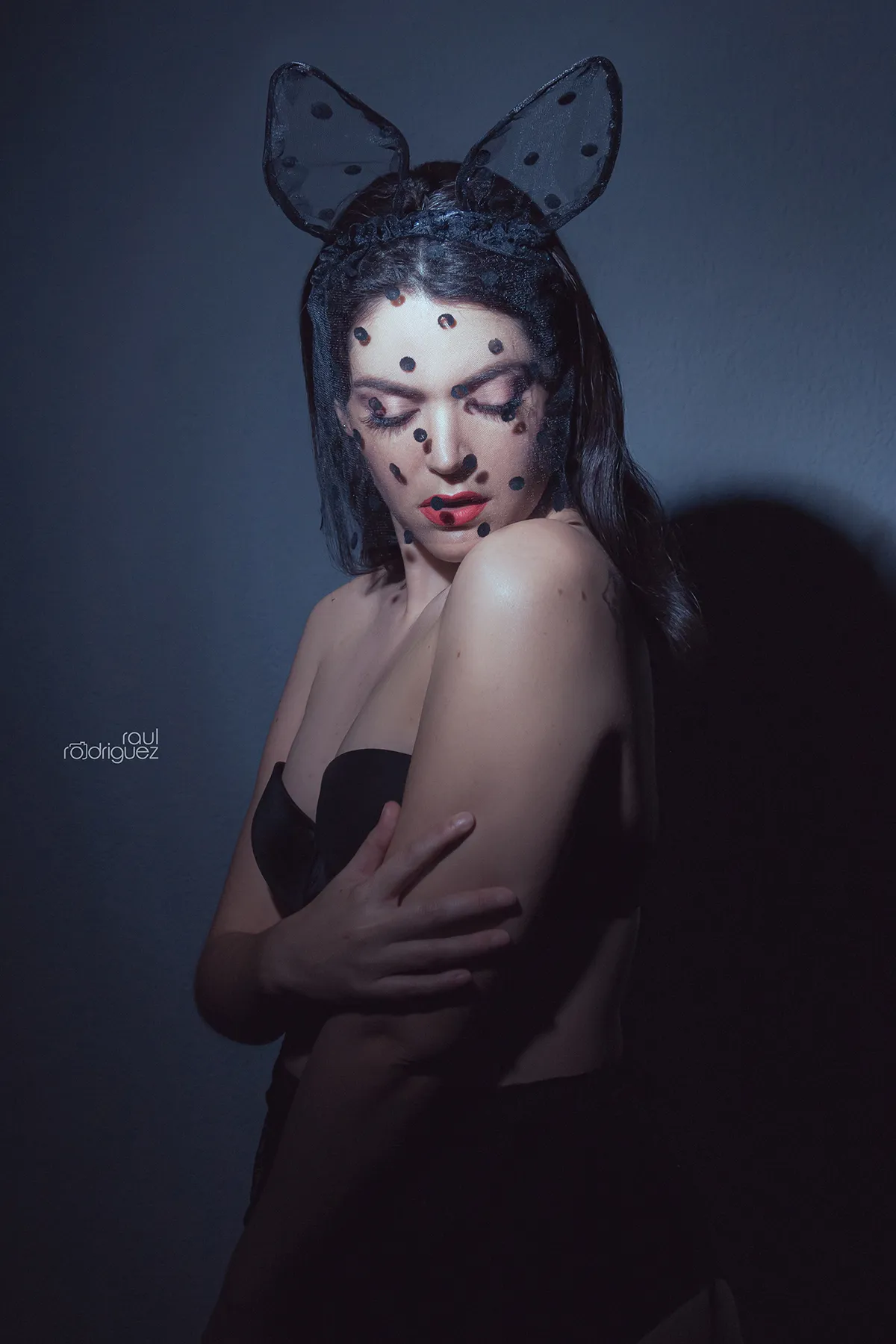 Retrato de mujer con orejas negras de conejo y velo de puntos, mirando hacia su hombro bajo una luz dirigida.