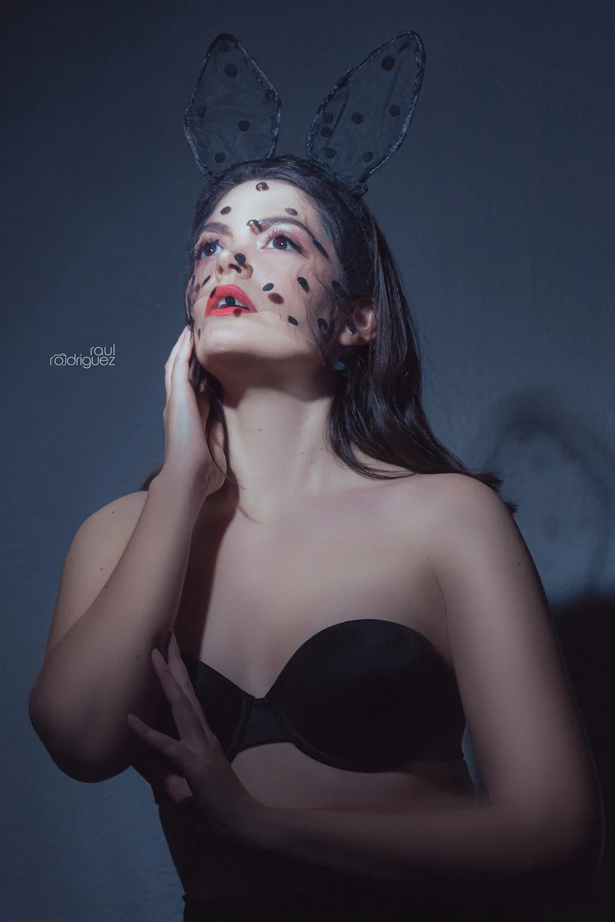Retrato de mujer con orejas negras de conejo y velo de puntos, mirando hacia arriba con una mano junto al cuello.