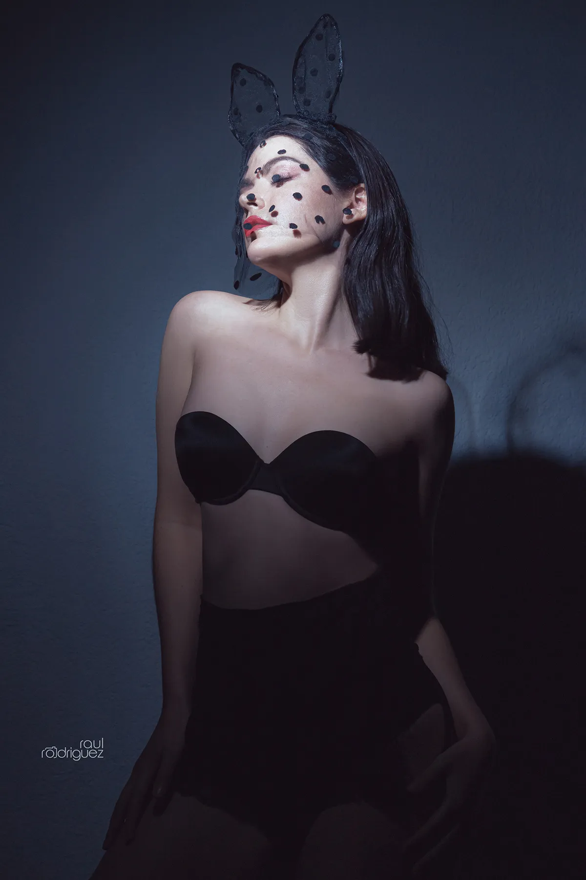 Retrato de mujer con orejas negras de conejo, rostro de perfil elevado y vestuario negro sobre un fondo oscuro.
