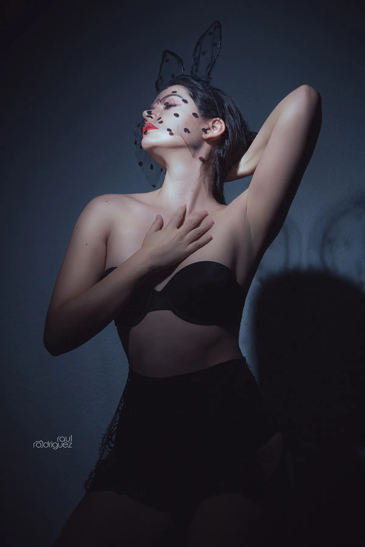 Retrato de mujer con orejas negras de conejo y velo de puntos, un brazo detrás de la cabeza y una mano sobre el pecho.