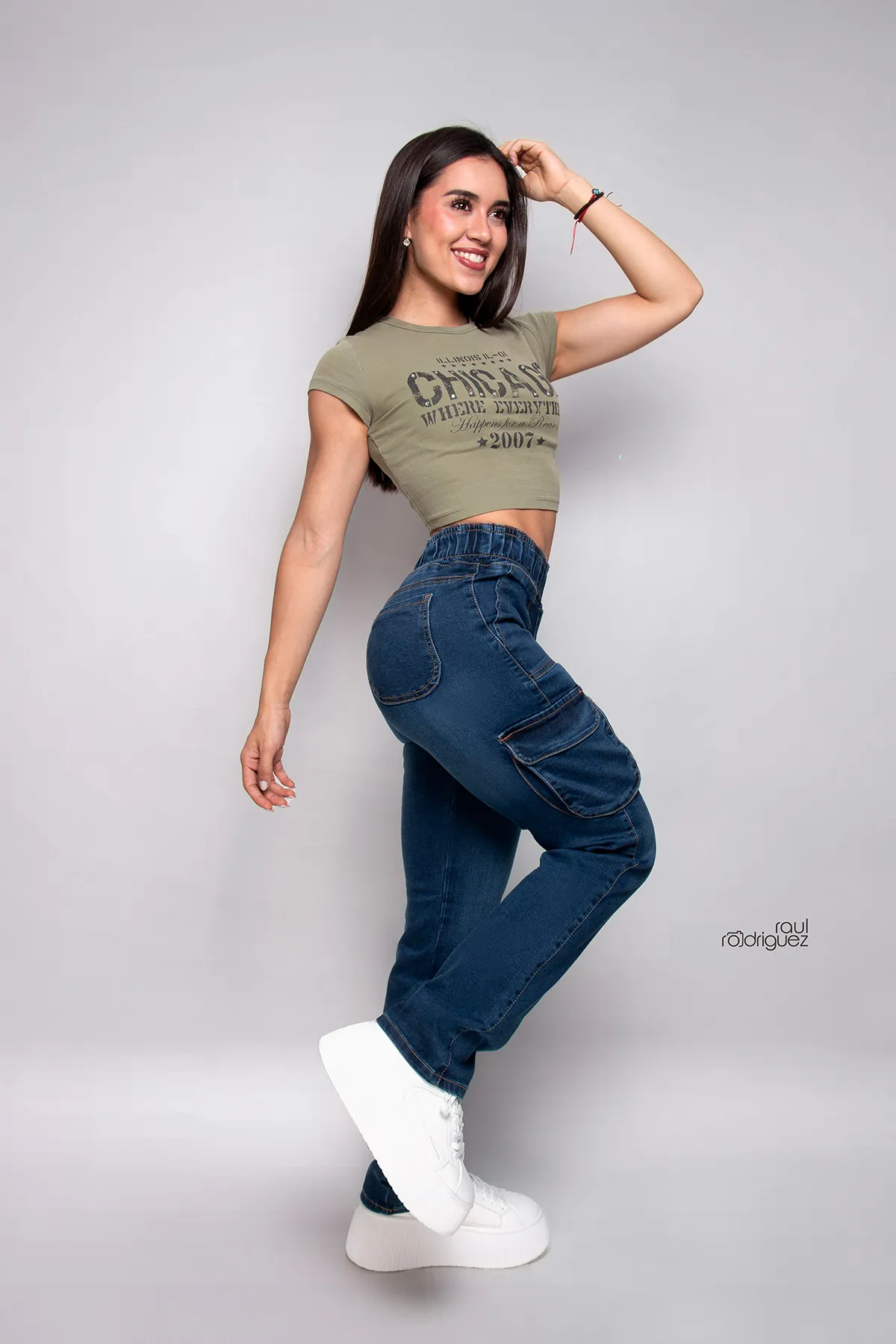 Mujer de cuerpo completo con pantalón cargo de mezclilla azul, camiseta corta verde y tenis blancos, posando de lado sobre un fondo gris claro.