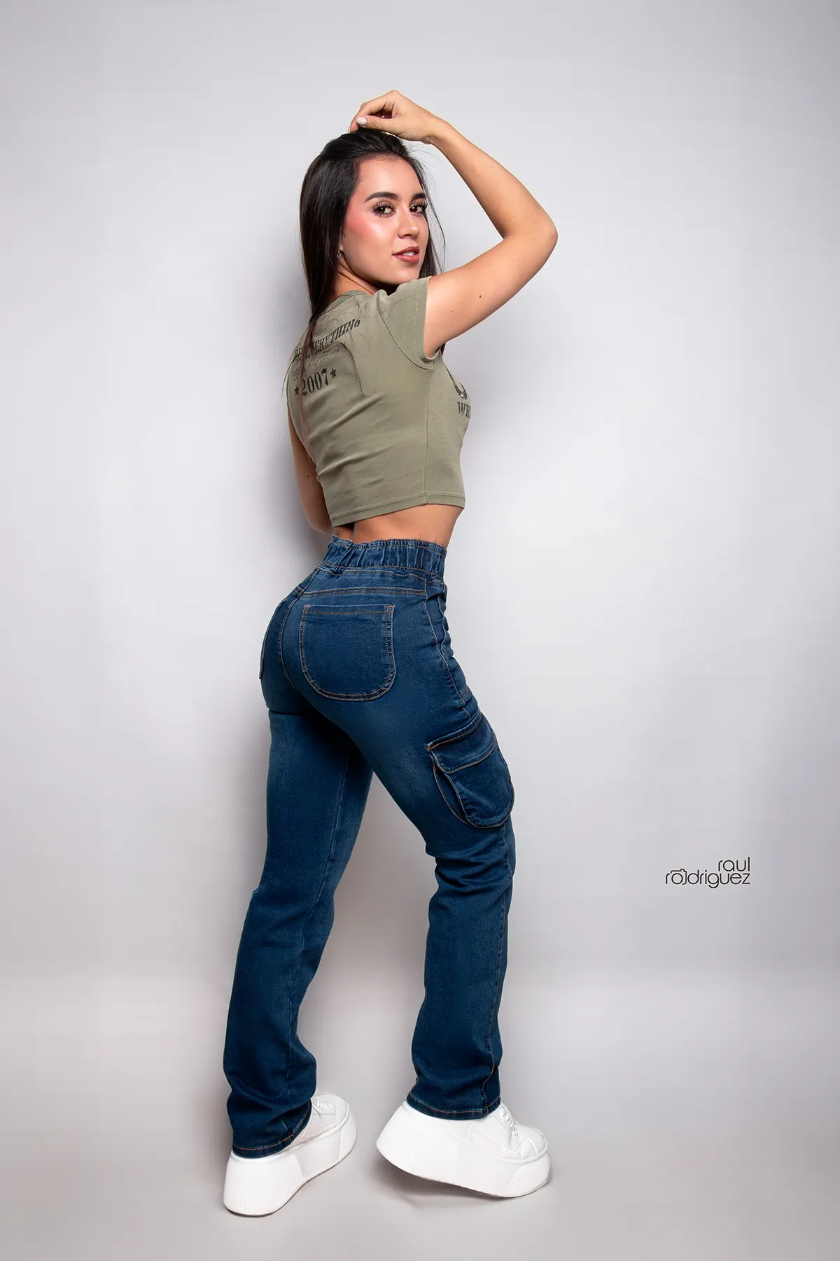 Mujer con pantalón cargo de mezclilla azul vista de espaldas y de lado, con una mano en el cabello y tenis blancos, sobre fondo gris claro.