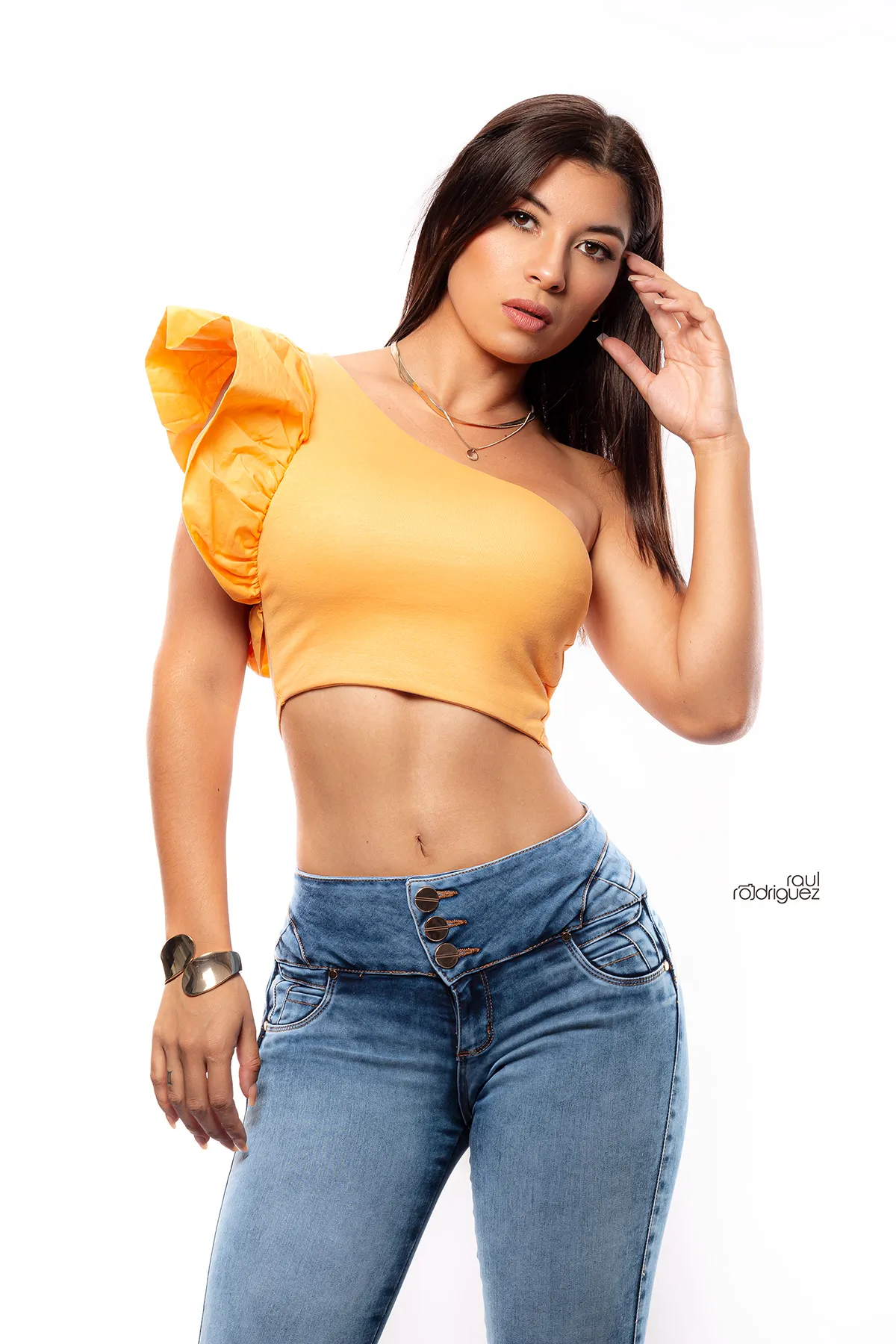 Mujer con blusa corta amarilla de un hombro y jeans azul claro posa con una mano junto al cabello frente a un fondo blanco.