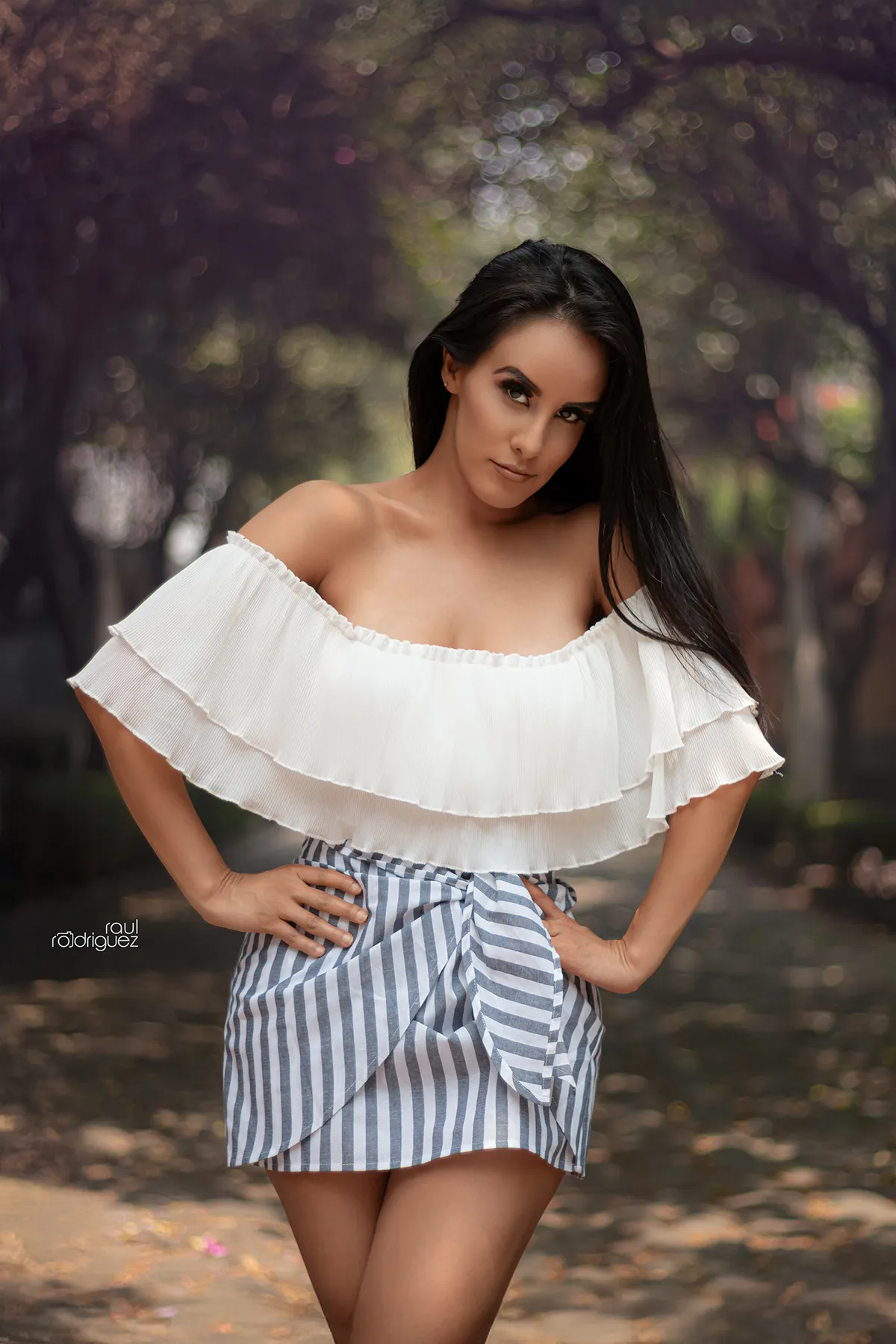 Mujer con blusa blanca de hombros descubiertos y falda corta de rayas posa en un sendero arbolado de Chimalistac.
