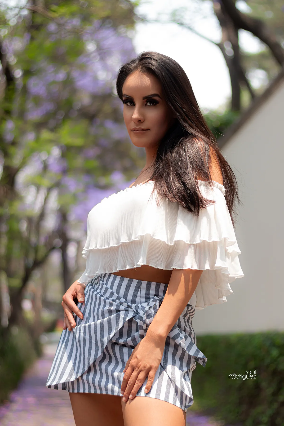 Mujer con blusa blanca de hombros descubiertos y falda de rayas posa al aire libre frente a un fondo de vegetación desenfocada.