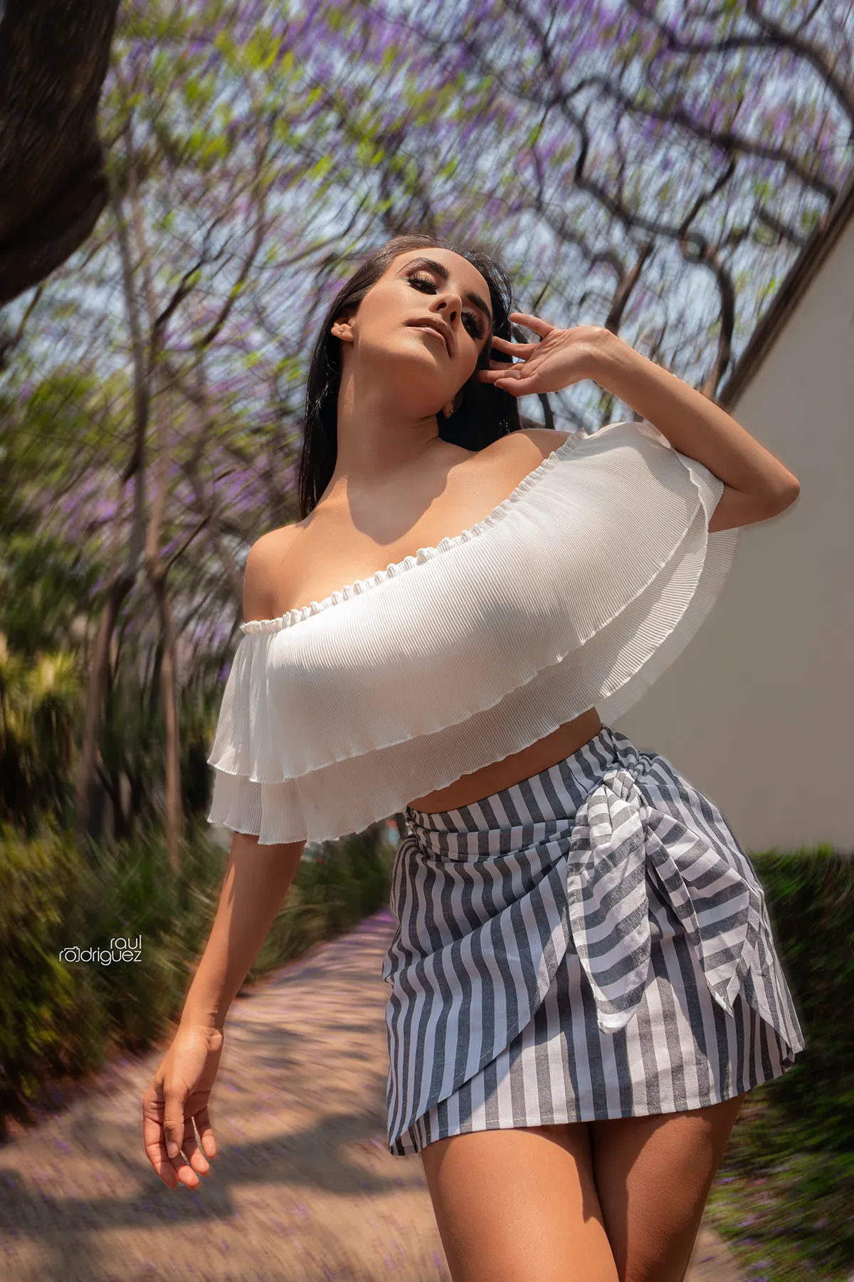 Mujer con blusa blanca de hombros descubiertos y falda de rayas posa en exteriores con árboles al fondo.