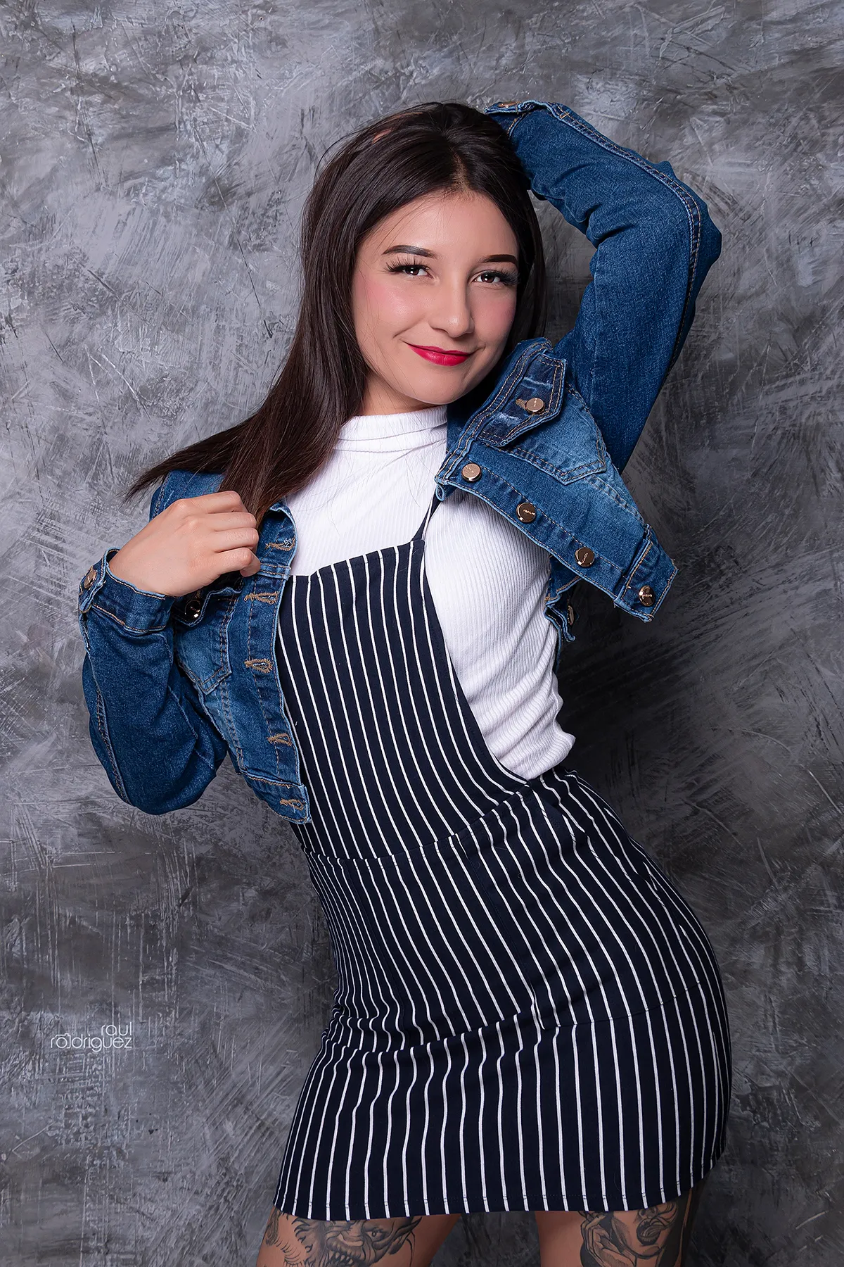Modelo con chamarra corta de mezclilla azul abierta, una mano en el cabello y otra sujetando la prenda, frente a un fondo gris texturizado.