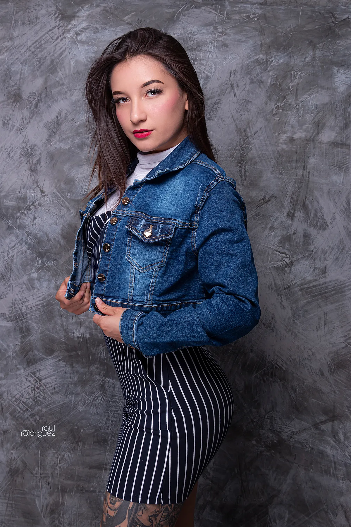 Modelo en pose de tres cuartos con chamarra corta de mezclilla azul abierta, bolsillos de pecho y botones metálicos, sobre fondo gris texturizado.