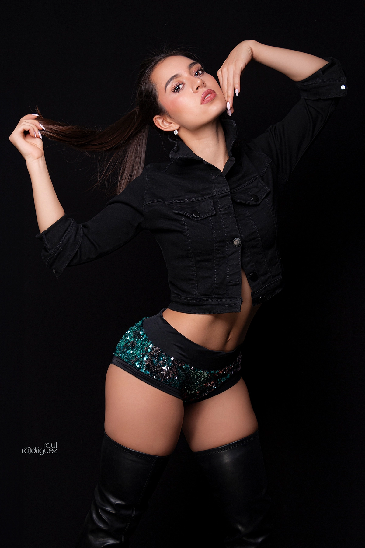 Mujer con chaqueta negra, shorts de lentejuelas y botas altas posa sobre fondo negro sosteniendo su cabello.