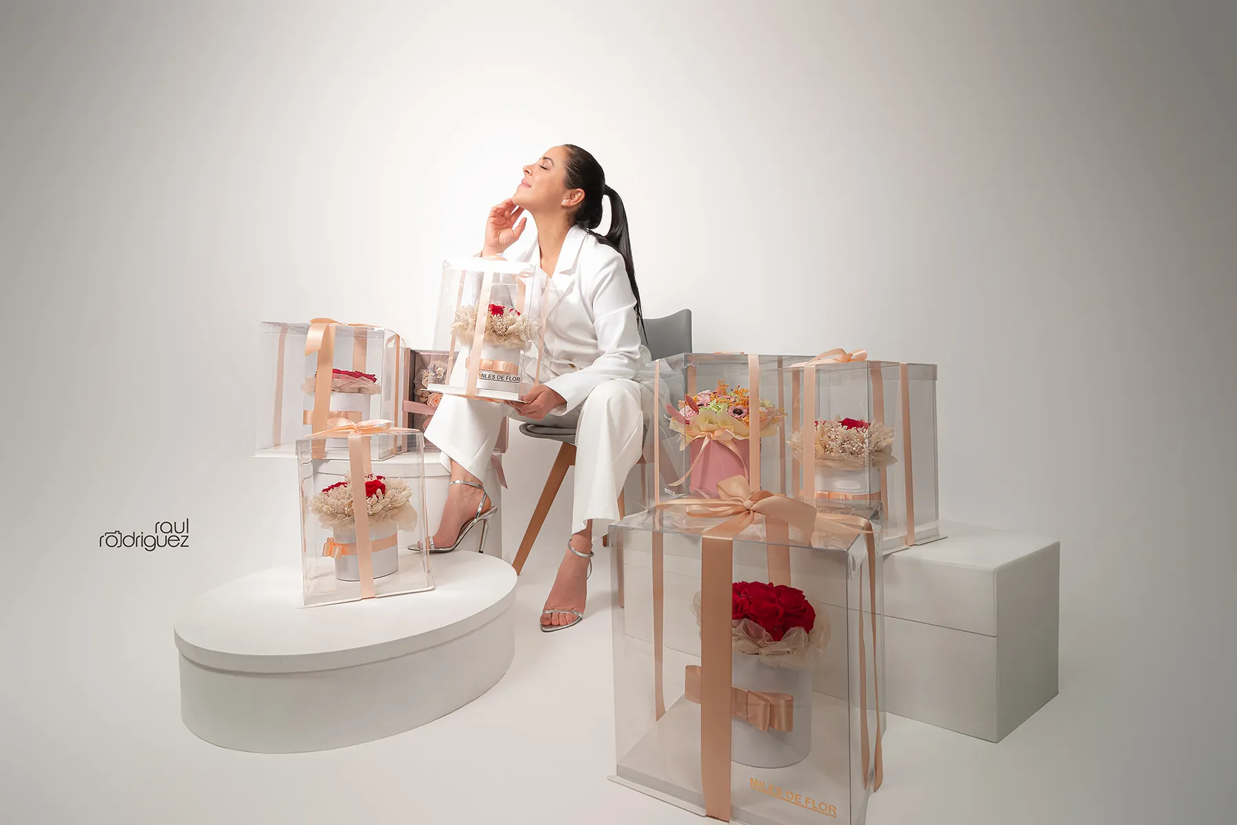 Mujer vestida de blanco sentada entre cajas transparentes con arreglos florales sobre fondo claro.